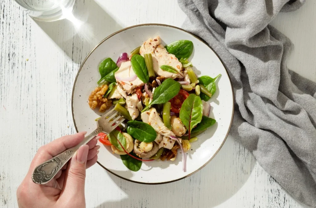 Hand holding a fork over a plate of keto chicken salad with spinach, walnuts, cherry tomatoes, green beans, onions, and white beans on a white wooden table
